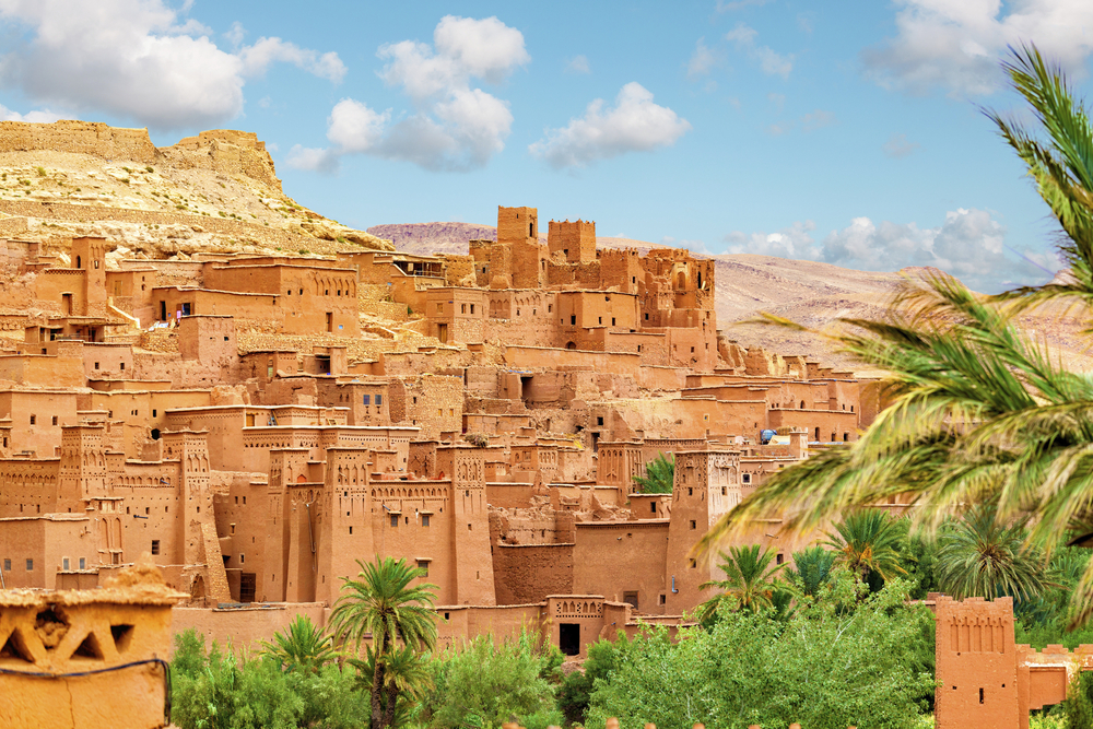 Dicht bebaute Lehmstadt Ait-Ben-Haddou mit gestaffelten, sandfarbenen Häusern und Türmen, umgeben von Palmen und karger Berglandschaft